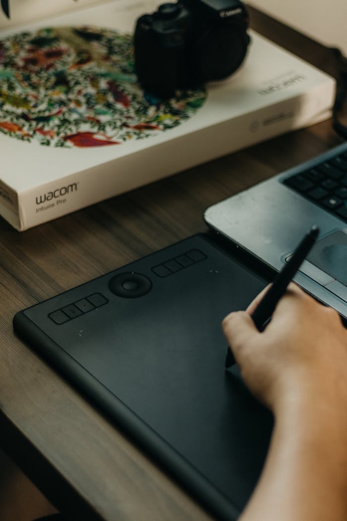 Close-up of a hand sketching with a stylus on a Wacom tablet beside a laptop on a desk.