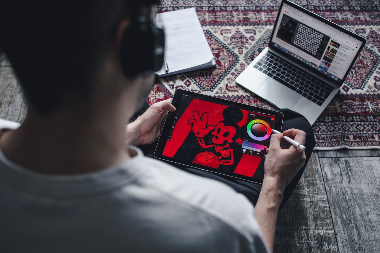 A person drawing on a tablet with a laptop nearby, showcasing digital art creation at home.